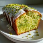 A slice cut from a moist pistachio bread loaf, topped with white glaze and chopped pistachios.