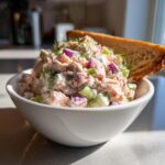 A close-up of creamy salmon salad mixed with celery and red onion, garnished with dill, served in a white bowl with toast.