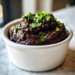A close-up of thick, dark black bean dip topped with fresh cilantro in a small white bowl.