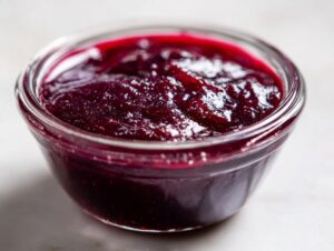 Close-up of thick, deep purple plum jam glistening in a small clear glass bowl.