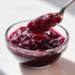 A spoonful of thick, dark red plum jam being lifted from a small glass bowl.