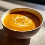 A close-up of vibrant orange roasted butternut squash soup, swirled with cream and black pepper.