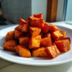 A close-up of beautifully caramelized, roasted sweet potatoes cubes piled high on a white plate.