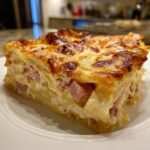A thick, square serving of scalloped potatoes and ham, showing layers of creamy potatoes and pink ham chunks under a browned, cheesy crust.