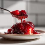 A spoonful of thick, glistening red christmas jam with whole cranberries being drizzled over a dessert on a white plate.