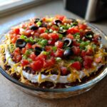 A colorful and fully loaded Seven layer dip in a clear glass bowl, topped with tomatoes, olives, and green onions.