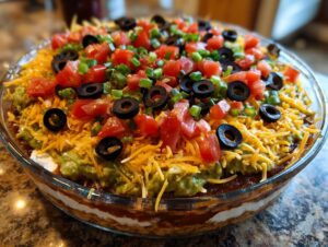A vibrant, fully assembled Seven layer dip topped with shredded cheese, diced tomatoes, black olives, and green onions.