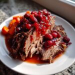 Tender, shredded cranberry pot roast topped with whole cranberries and served alongside cooked carrots on a white plate.