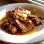 Close-up of tender, shredded Mississippi pot roast served in a white bowl with rich juices and two pats of melting butter.