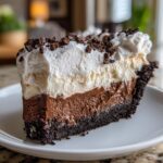 A decadent slice of mud pie featuring a dark crust, two layers of chocolate filling, and a thick layer of whipped cream topped with chocolate shavings.