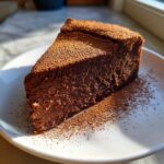 A rich, dense slice of flourless chocolate torte dusted heavily with cocoa powder, served on a white plate.