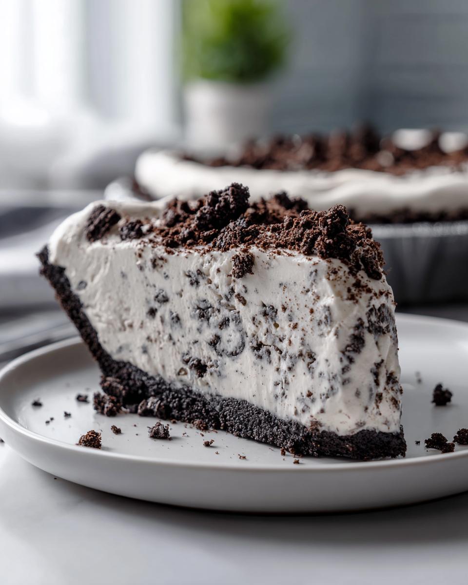 A thick slice of no-bake Oreo pie featuring a dark crust, creamy filling with cookie chunks, and crumb topping.