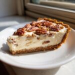 A close-up slice of rich pecan cream pie with a thick graham cracker crust and whole pecans on top.