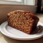 Close-up of a thick slice of moist Protein banana bread showing its dense, brown crumb texture on a white plate.