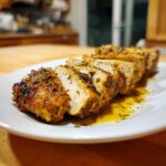 Close-up of sliced, herb-crusted greek chicken breasts resting in pan juices on a white plate.