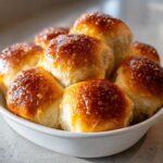 A close-up of several golden-brown, soft buttery rolls piled high in a white serving dish.