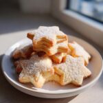 A stack of golden, star-shaped butter cookie treats dusted generously with powdered sugar on a white plate.