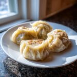 Four steamed Traditional Chinese Dumplings (likely Shumai style) served on a white plate near a window.