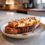 A slice of charred sweet potato toast topped with yogurt, peanut butter, chopped nuts, and drizzled with syrup.