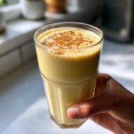 Hand holding a tall glass filled with a thick, yellow banana smoothie, dusted with cinnamon.