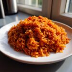 A mound of vibrant, orange-red jollof rice served on a white plate, brightly lit by natural window light.