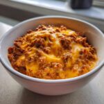 Close-up of a white bowl filled with rich chili cheese dip topped with melted, gooey cheddar cheese.