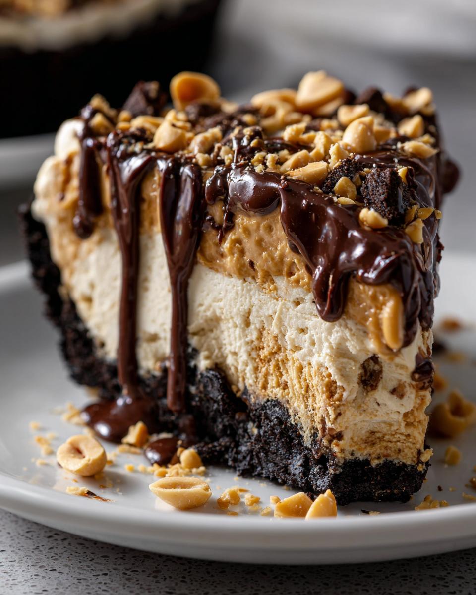 Close-up of a decadent slice of ice cream pie with peanut butter and vanilla layers, topped with chocolate fudge and chopped peanuts.