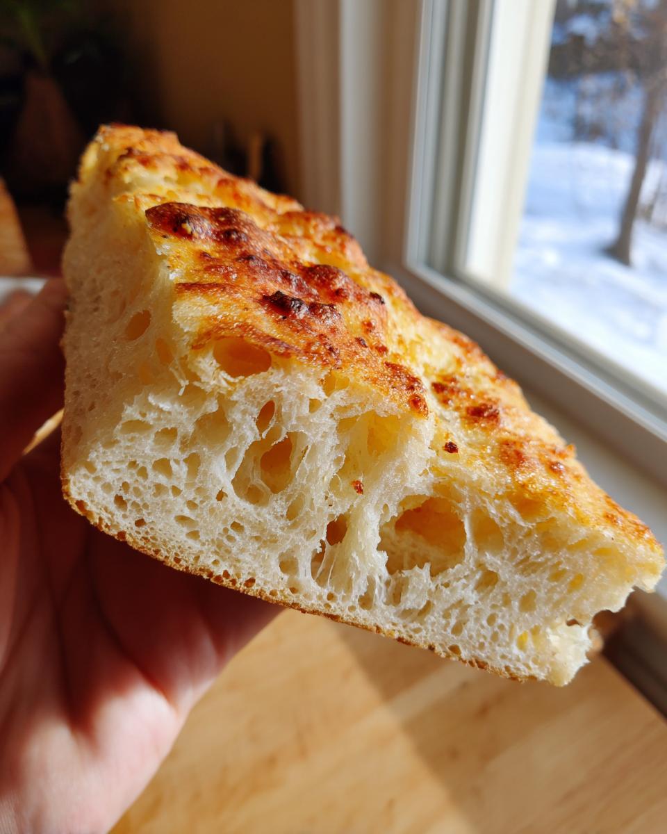 A hand holding a thick slice of airy, golden-brown keto pizza crust showing large air pockets.