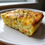 A close-up of a square slice of baked egg bake casserole with visible carrots and chives on top.