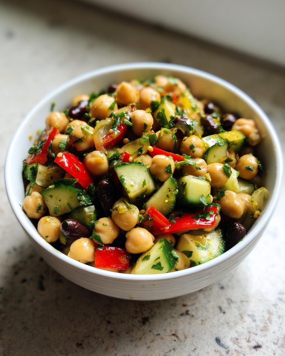 A close-up of a vibrant bowl of fresh bean salad featuring chickpeas, cucumber, red pepper, and herbs.