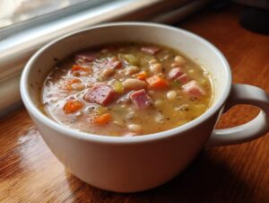 Close-up of a white mug filled with thick, comforting bean soup containing ham chunks and diced carrots.