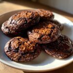 A stack of rich, dark chocolate brownie cookies featuring classic shiny, crinkly tops, resting on a white plate.