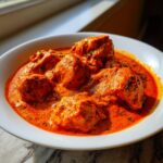 Close-up of tender chicken pieces swimming in a vibrant orange, creamy sauce of butter chicken, garnished with herbs.