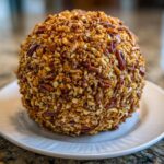 A large, perfectly round cheese ball recipe coated entirely in chopped pecans, resting on a white plate.