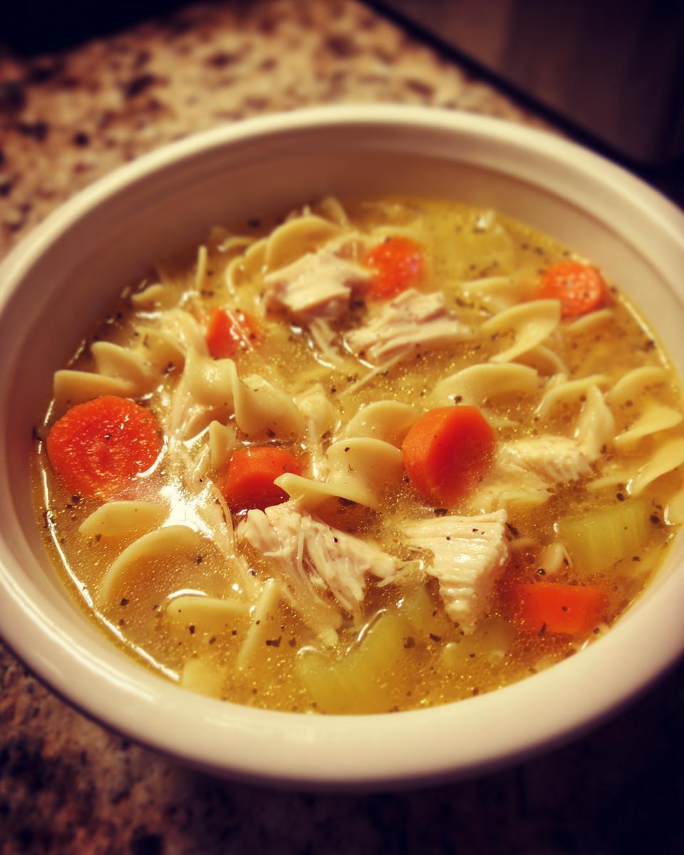 Close-up of a white bowl filled with steaming chicken noodle soup featuring egg noodles, shredded chicken, and bright orange carrots.