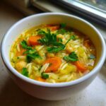Close-up of a white bowl filled with vibrant yellow chickpea soup, featuring carrots, orzo pasta, and fresh parsley garnish.