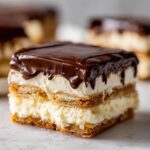Close-up of a square slice of eclair cake showing layers of cream filling, graham crackers, and thick chocolate ganache topping.