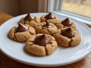 A white plate holds several freshly baked kiss cookies topped with melted milk chocolate Hershey's Kisses.