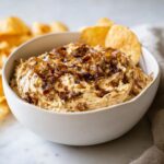 A creamy bowl of homemade onion dip topped with caramelized onions, served with potato chips.
