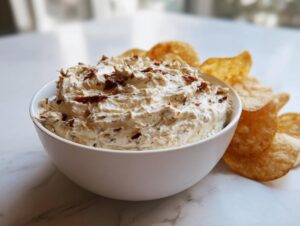 A bowl of creamy, textured onion dip topped with crispy bits, served with potato chips.