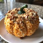 A festive pumpkin cheese ball coated in orange cheese and chopped pecans, featuring a real pumpkin stem.