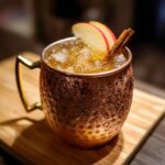 A frosty copper mug filled with an apple cider Moscow mule cocktail, garnished with apple slices and a cinnamon stick.