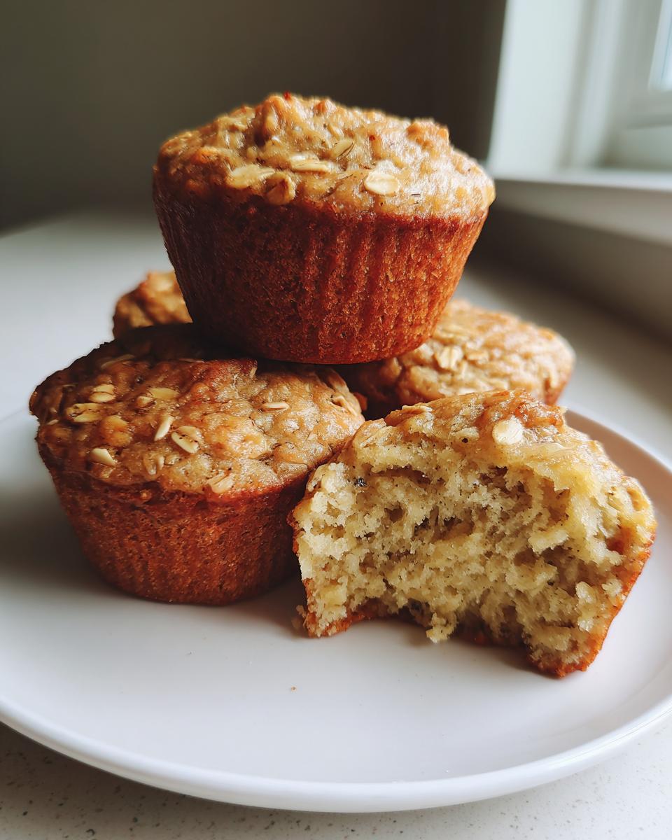 A stack of golden brown banana oatmeal muffins, one partially eaten to show the moist interior.