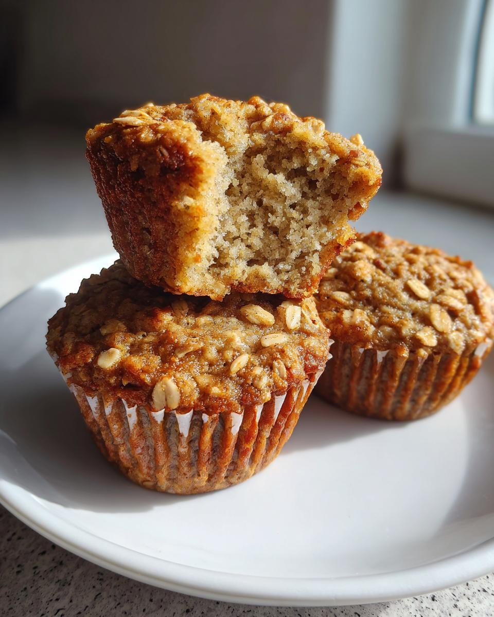 Three delicious banana oatmeal muffins stacked on a white plate, one with a bite taken out showing the moist interior.