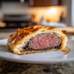 A cross-section slice of beef wellington recipe showing medium-rare beef inside flaky, golden puff pastry.