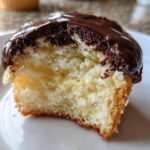 Close-up of a Boston cream pie cupcakes with a bite taken out, showing vanilla cake, pastry cream filling, and chocolate glaze.