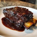 A single serving of rich, dark braised short ribs covered in glossy sauce, resting on a white plate.
