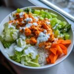 A vibrant buffalo chicken bowl featuring shredded orange buffalo chicken, white rice, lettuce, celery, carrots, and ranch dressing.