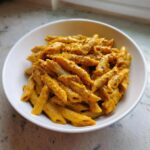 A white bowl filled with penne pasta coated in a rich, orange butternut squash pasta sauce and sprinkled with black pepper.