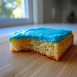 Close-up of a square of yellow cake topped with vibrant blue cookie icing recipe, resting on a wooden cutting board.